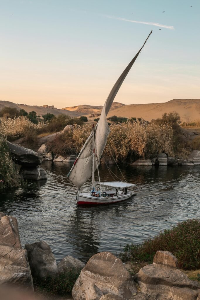 A serene sailboat glides along a river surrounded by rocks and hills during golden sunset light.